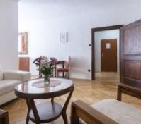 Útulný obývací pokoj s bílou koženou pohovkou, kulatým dřevěným konferenčním stolkem s vázou s květinami a dřevěnými podlahami. Na chodbu vedou dřevěné dveře s malým stolem a židlemi v pozadí.