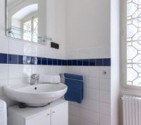 Bílý dřez na úložné skříňce s velkým zrcadlem a skleněnou poličkou nahoře. Modré dekorativní dlaždice a věšák na ručníky dodávají barvu. Okno s dekorativními mřížkami propouští přirozené světlo.